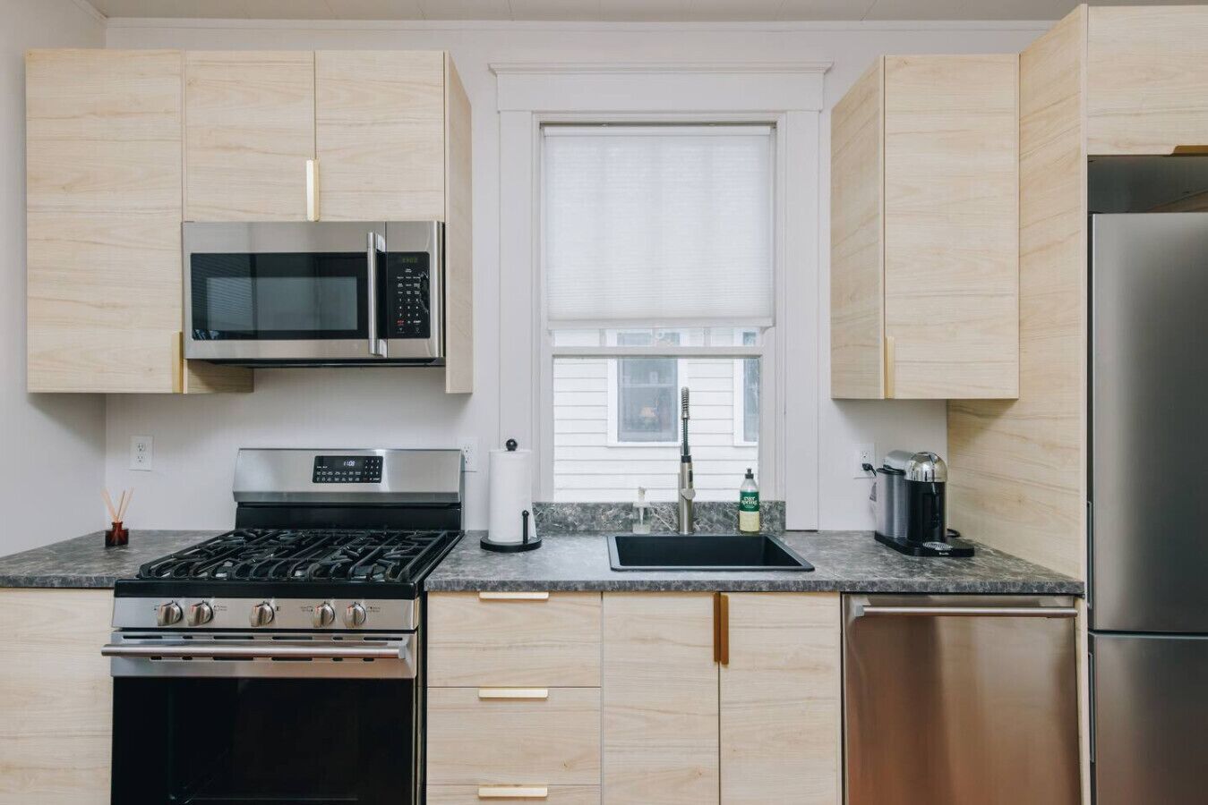 Una cocina moderna con armarios de madera clara, electrodomésticos de acero inoxidable, incluyendo un microondas, estufa y refrigerador, y una ventana sobre el fregadero.
