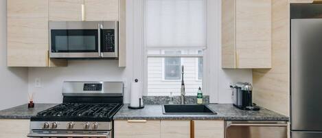 Una cocina moderna con armarios de madera clara, electrodomésticos de acero inoxidable, incluyendo un microondas, estufa y refrigerador, y una ventana sobre el fregadero.