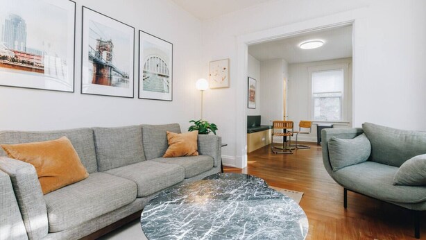 A modern living room with a grey sofa, a round marble coffee table, and a dining area with a wooden table and chairs. - Hyde Park 3BR Home with Fenced Backyard (Cincinnati)