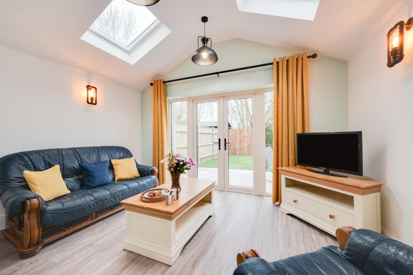 Un salon moderne avec un canapé en cuir, une table basse, un téléviseur posé sur un meuble en bois et une porte-fenêtre coulissante donnant sur un jardin.
