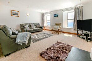 A modern living room with a sofa, armchair, and a flat-screen TV. - 3 Bed in Porthleven (oc-atlant) (Porthleven)