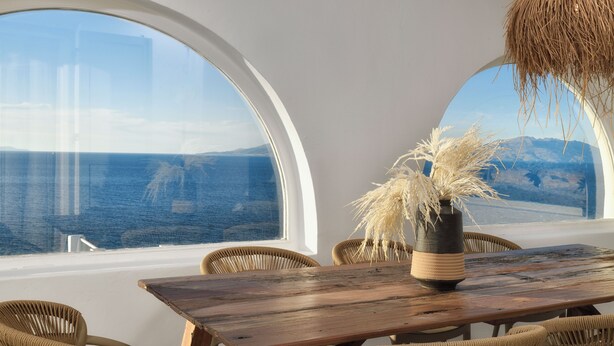 Ein Essbereich mit einem Holztisch, Korbstühlen, einer Vase mit getrockneten Pflanzen und Blick auf das Meer durch große Rundbogenfenster.