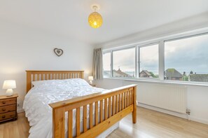 A bedroom with a wooden bed, a nightstand with a lamp, a large window with a view of houses, and a ceiling light. - 2 Bed in Broadstairs (oc-th019) (Broadstairs)