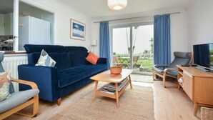 A modern living room with a blue sofa, a wooden coffee table, a TV, and a dining area with a table and chairs. - 3 Bed in Perranuthnoe (oc-4withe) (Perranuthnoe)