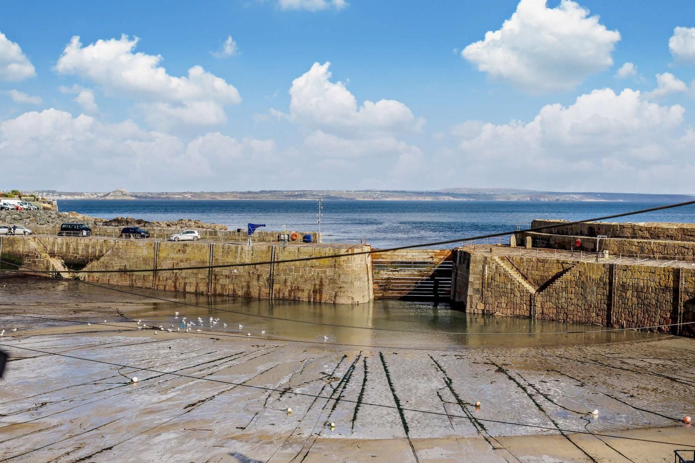 Une jetée côtière avec un escalier en pierre descendant vers l’eau, un ciel dégagé et une vue sur la mer et la terre au loin.