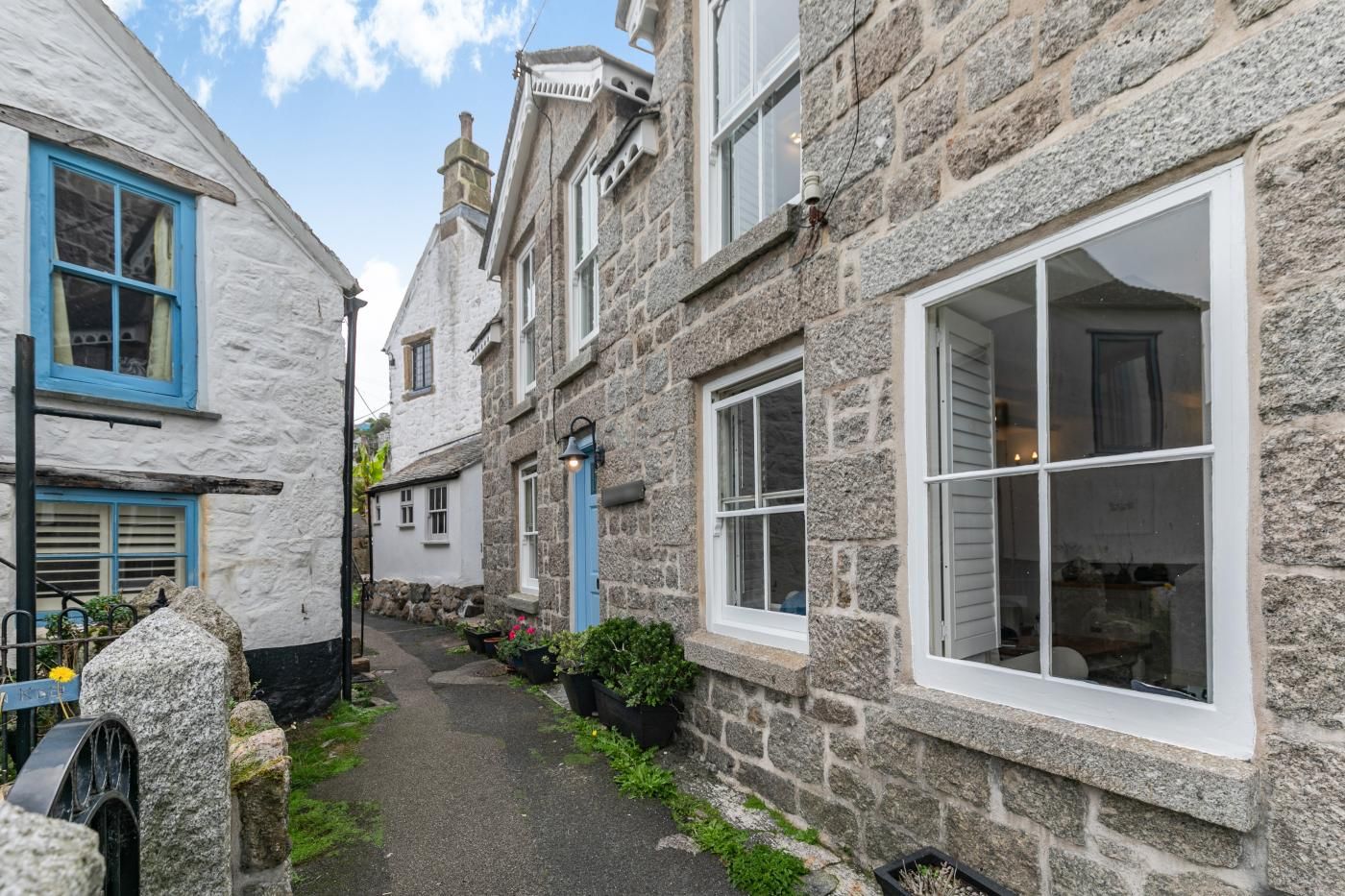 A narrow street with stone buildings, some with blue doors and windows, and a small pathway with greenery.