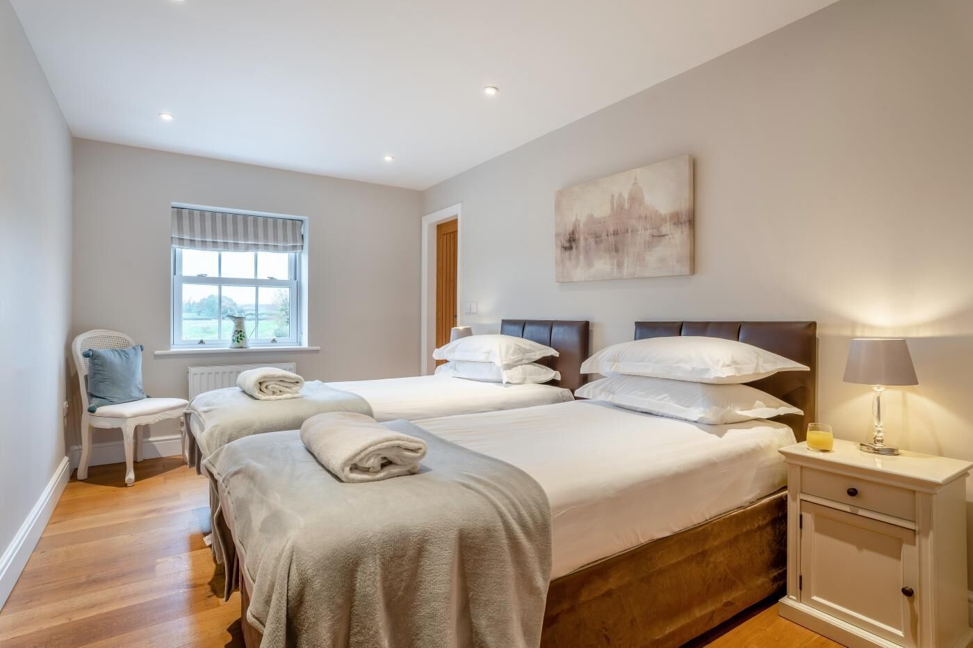 A hotel room with two beds, a chair, a window, a painting, and a bedside table with a lamp.