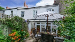 A patio with a table set for dining, surrounded by greenery and a conservatory. - 2 Bed in Mousehole (oc-threeb) (Mousehole)