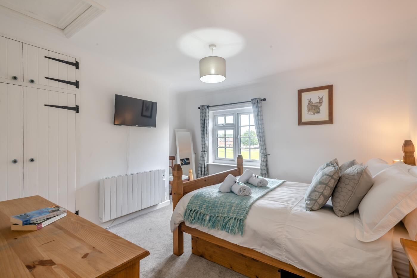 A bedroom with a bed, a wooden table, a TV, a window with curtains, and a radiator.