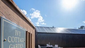 Een bordje voor 'Ivy Cottage Garden' met een houten hek, een gazon en een gebouw met een metalen dak op de achtergrond.