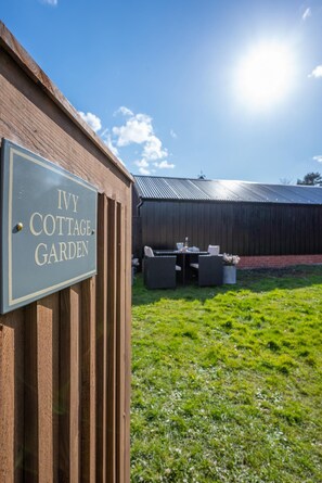 A sign for 'Ivy Cottage Garden' with a wooden fence, a grassy area, and a metal-roofed building in the background. - 2 Bed in Aldborough (oc-2257) (Aldborough)