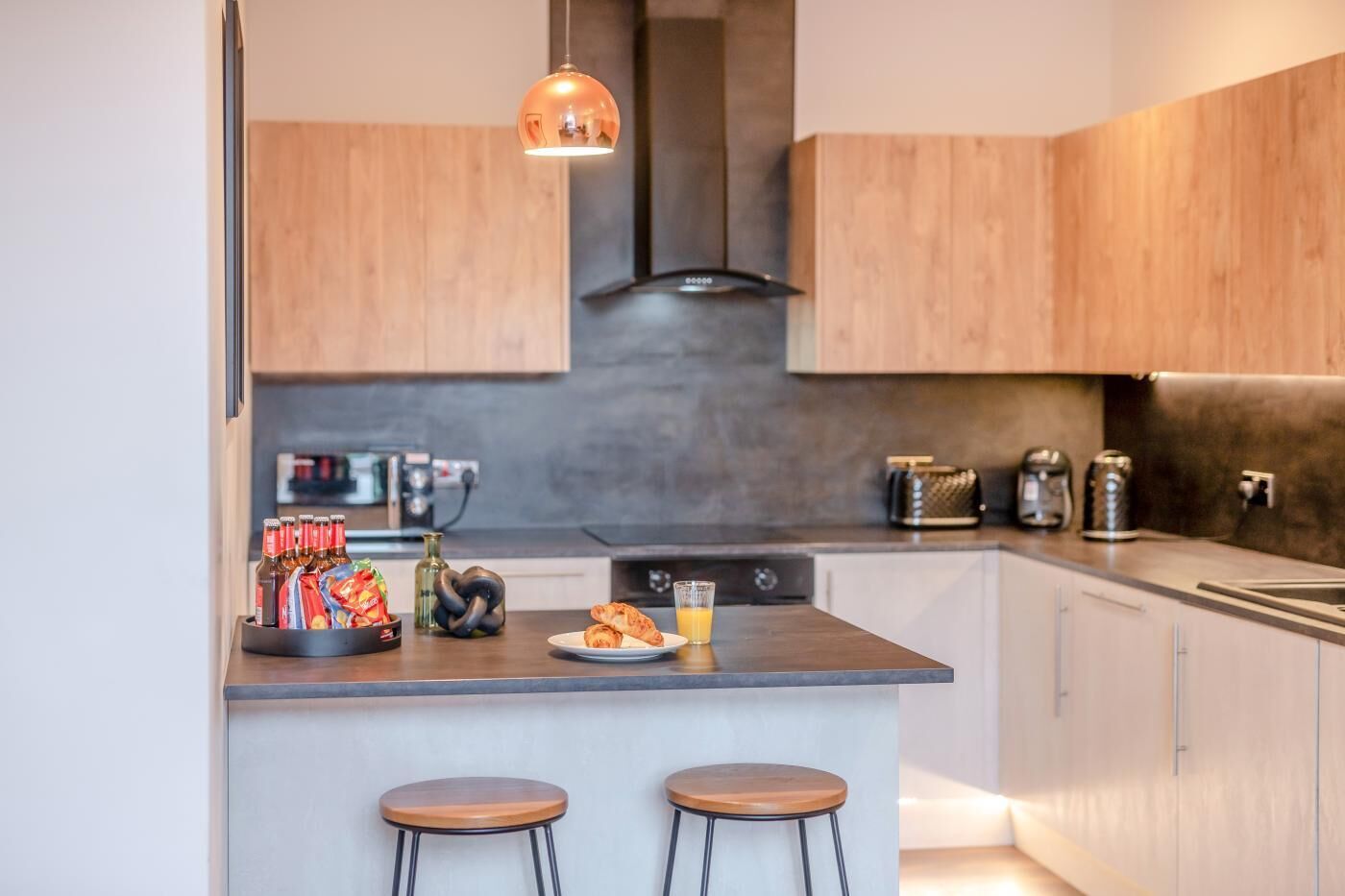 Une cuisine moderne avec des meubles en bois, un îlot central doté de tabourets, un luminaire suspendu et divers appareils de cuisine.