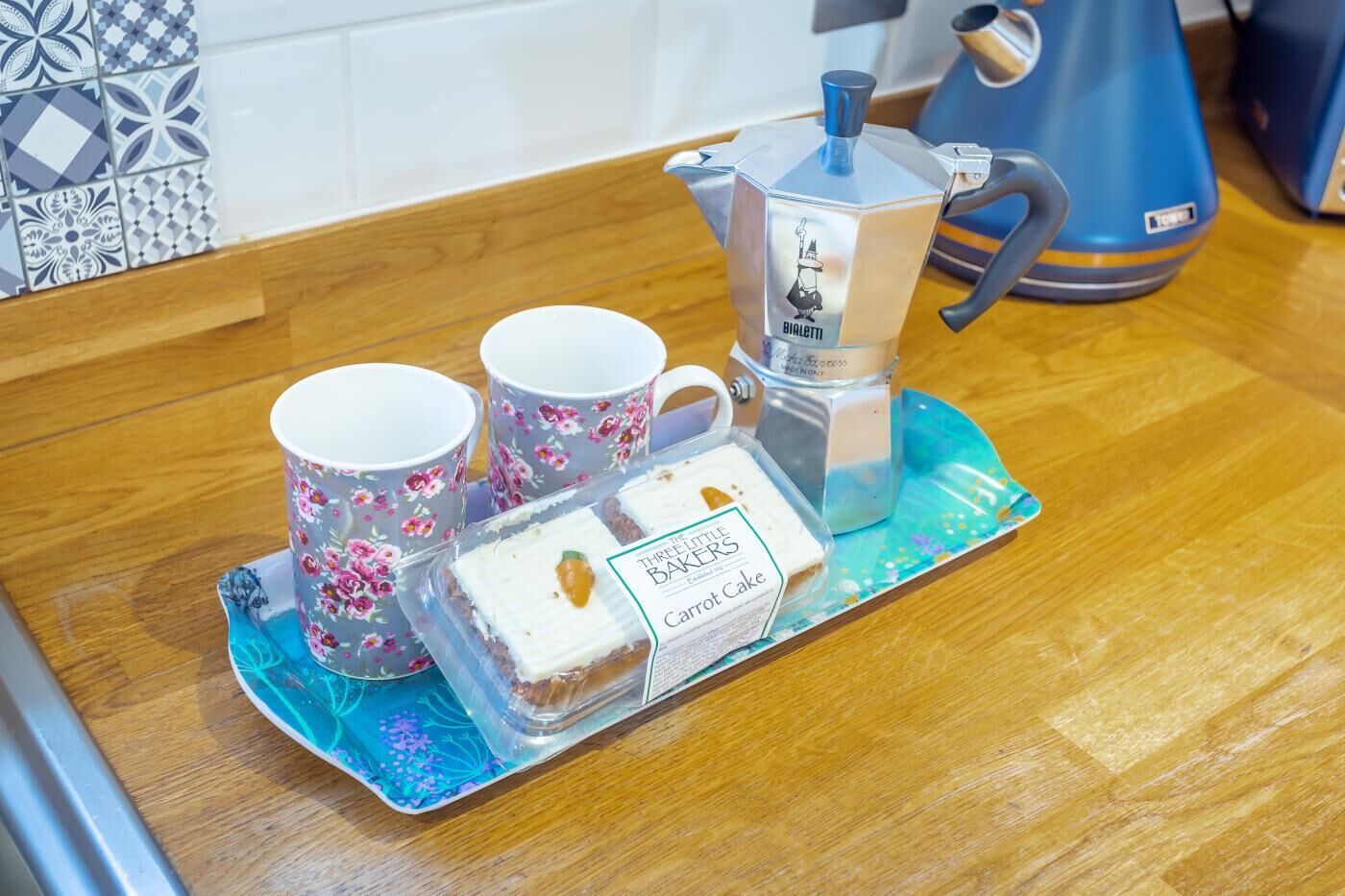 Un comptoir de cuisine avec une machine à café, deux tasses ornées de motifs floraux et un plateau contenant un gâteau et une dosette de café.