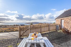 A wooden table with breakfast items, a stone building, and a view of the sea and hills. - 2 Bed in Big Sand (oc-d33686) (Big Sand)