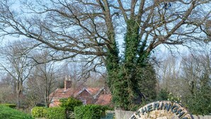 Un paysage rural avec, au premier plan, une sculpture circulaire en métal portant l’inscription « LETHERINGHAM », une route qui tourne vers la gauche et, au loin, une maison avec un toit rouge.