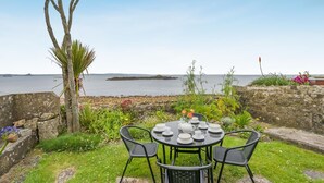 A seaside patio with a table set for four, surrounded by greenery and a view of the sea.