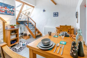 A dining area with a wooden table set for two, a staircase with a blue net, and a view of the outside. - 1 Bed in Porthleven (oc-smuggl) (Porthleven)