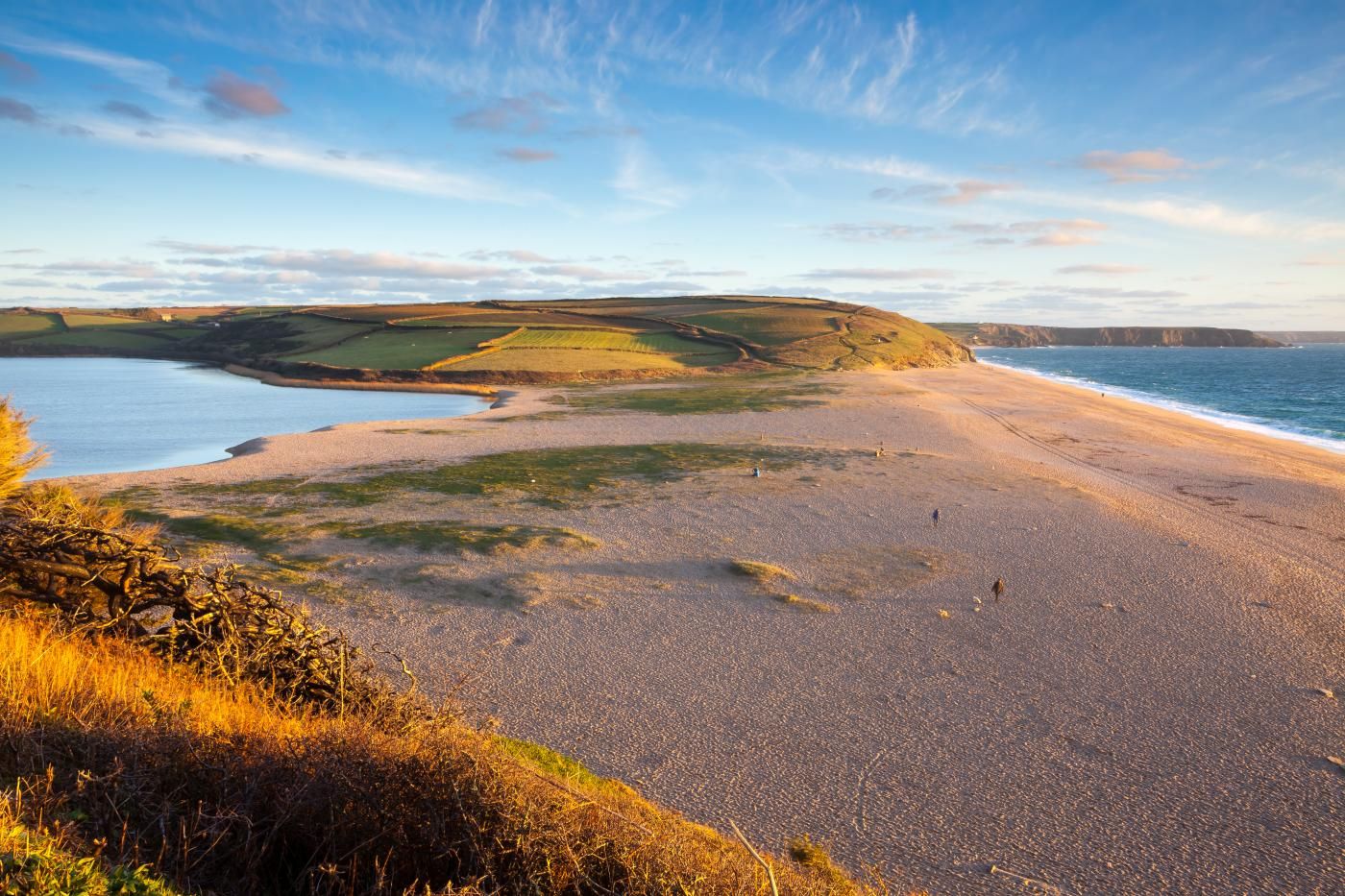 A coastal landscape with a pebbly beach, grassy areas, and rolling hills in the background.