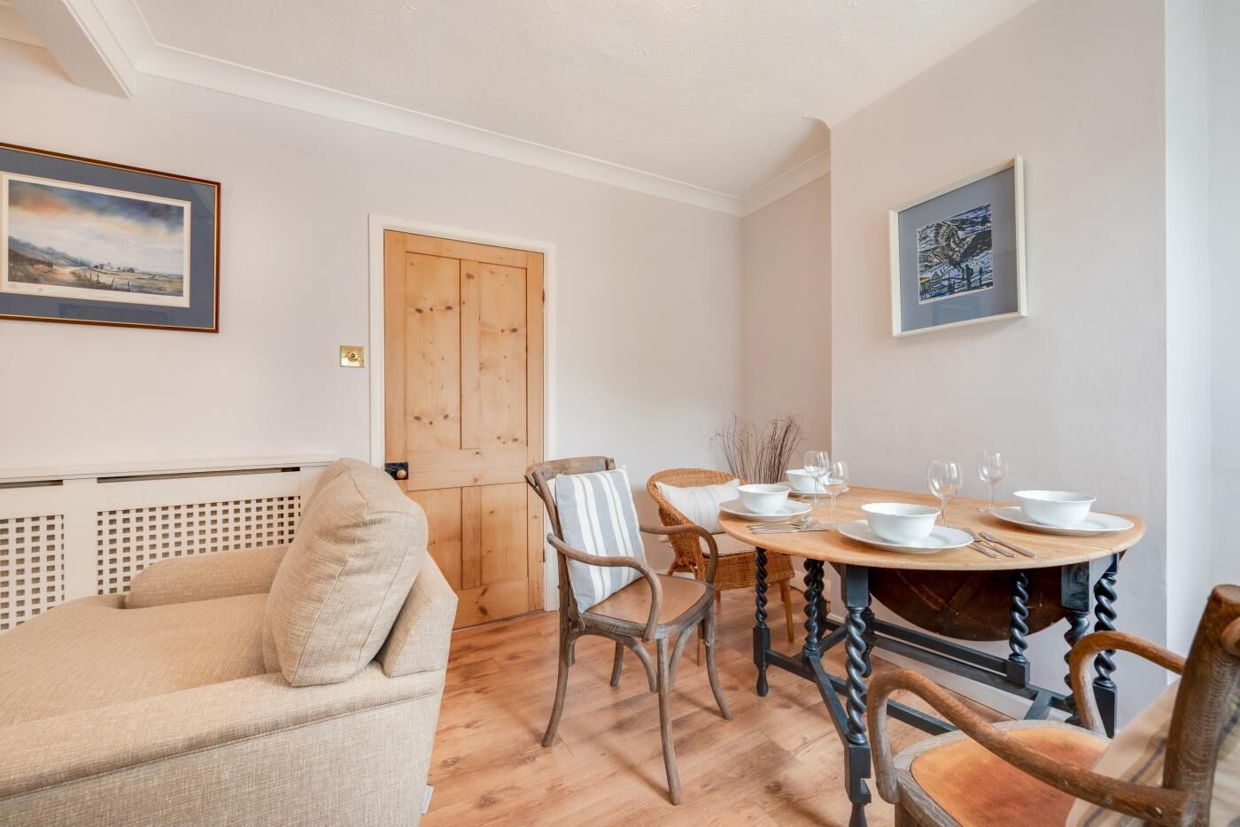 A cozy living room with a beige sofa, a wooden dining table set for four, and a wooden door.