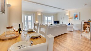 A dining area with a wooden table set for a meal, a television in the background, and a view of the sea through large windows. - 2 Bed in Porthleven (oc-marin2) (Porthleven)