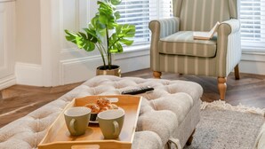 Una sala acogedora con un sillón a rayas, una planta en maceta, una bandeja con productos horneados y tazas, y un control remoto sobre un otoman tejido.