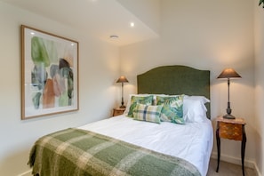 A neatly made bed with a green blanket, two bedside lamps, and a framed abstract painting on the wall.