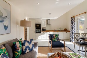 A modern living room with a sofa and armchair, a dining table with chairs, a kitchen with white cabinets, and a large window with curtains.