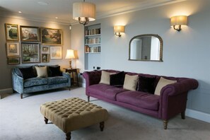 Ein Wohnzimmer mit einem blauen Sofa, einem violetten Sofa und einem beigefarbenen Hocker. An den Wänden sind Lampen angebracht und ein großes Spiegel ist zu sehen.