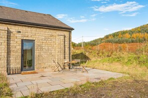 A stone building with a door and a window, a patio with a table and chairs, and a hilly landscape in the background.