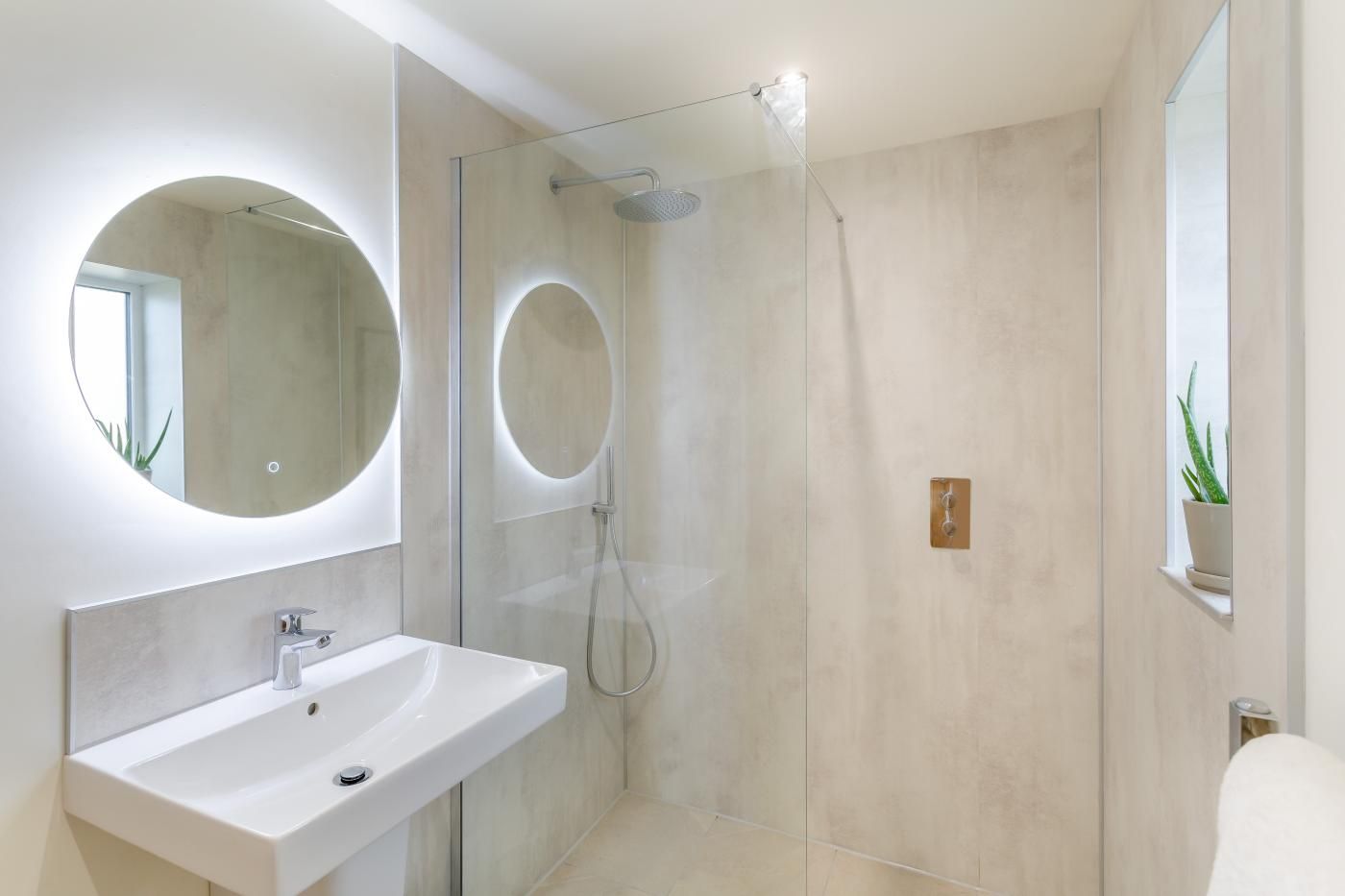 Un baño moderno con ducha envuelta en vidrio, un espejo redondo y un lavamanos blanco.