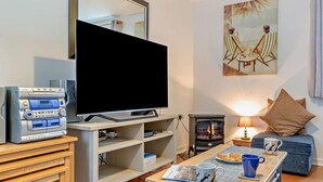 Uma sala de estar com televisão de tela plana, lareira a lenha, sofá, mesa de centro com canecas e um quadro na parede.