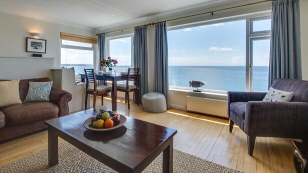 A spacious living room with a wooden floor, a brown leather sofa, a striped armchair, and a dining table with chairs. A large window offers a view of the sea. - 2 Bed in Porthleven (oc-lampho) (Porthleven)
