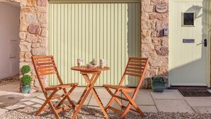 Un bâtiment en pierre avec une porte verte, deux chaises en bois, une petite table ronde et des plantes en pot.
