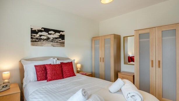 Un lit bien fait, avec des oreillers blancs et rouges, une lampe de chevet et un téléphone sur une table de chevet en bois. Il y a une grande armoire avec un miroir et un tableau encadré accroché au mur au-dessus du lit.