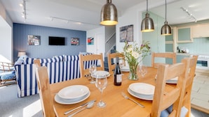 A dining area with a wooden table set for two, a blue-striped sofa, and hanging pendant lights. - 3 Bed in Porthleven (oc-oceavi) (Porthleven)