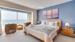 A bedroom with a large bed, a view of the sea, wicker chairs, and a sunset painting on the wall. - 3 Bed in Porthleven (oc-oceavi) (Porthleven)