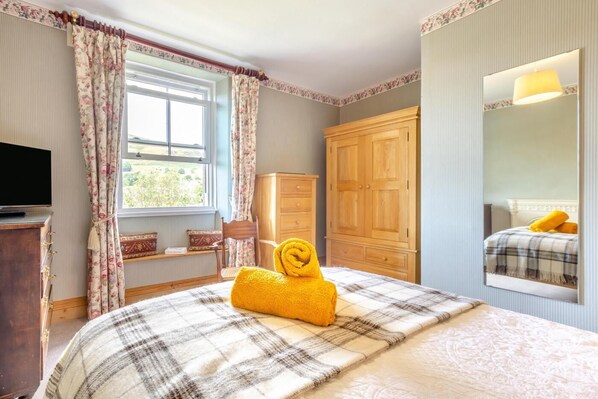 A bedroom with a bed, a wooden wardrobe, a television on a stand, and a window with curtains.