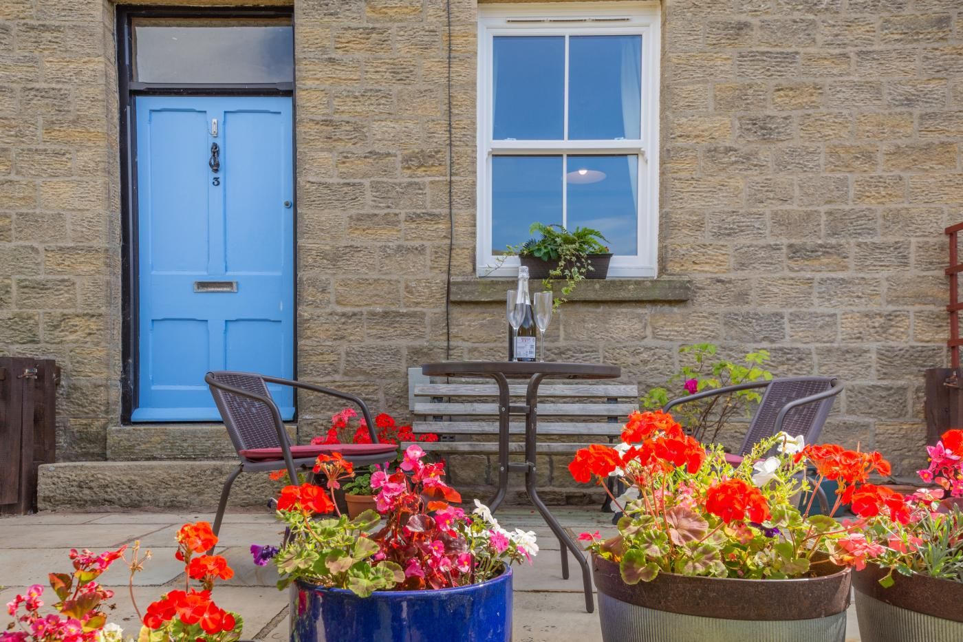 A stone building with a blue door, a window with a hanging plant, and outdoor seating with chairs and tables.