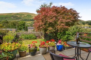 A patio with potted plants, a table, and chairs, overlooking a scenic landscape with hills and a stone wall.
