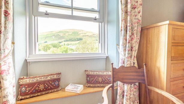 Una habitación con ventana que da a un paisaje verde, una silla de madera, un armario de madera y un banco con cojines.