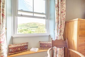 Ein Zimmer mit Fensterblick auf eine grüne Landschaft, ein Holzsessel, ein Holzschrank und eine Bank mit Kissen.