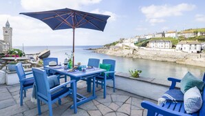 A seaside patio with blue furniture, a table set for dining, and a view of the sea and a historic tower. - 3 Bed in Porthleven (oc-12bayv) (Porthleven)