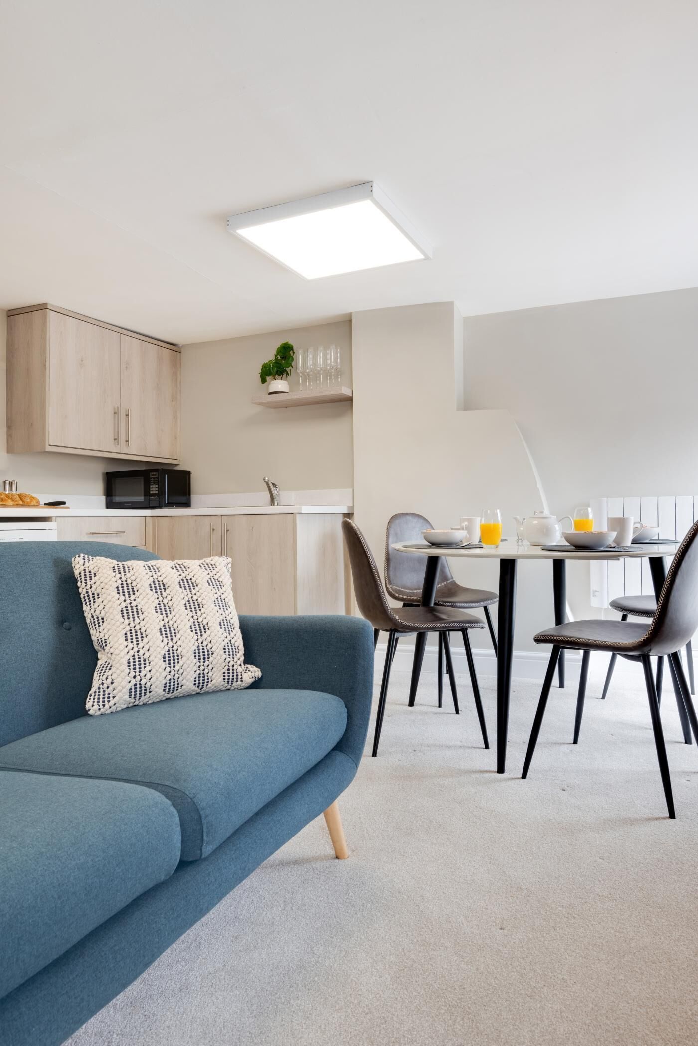 Un salon moderne avec un canapé bleu, une table à manger avec des chaises, un coin cuisine doté de placards et d’un four à micro-ondes, et un luminaire au plafond.