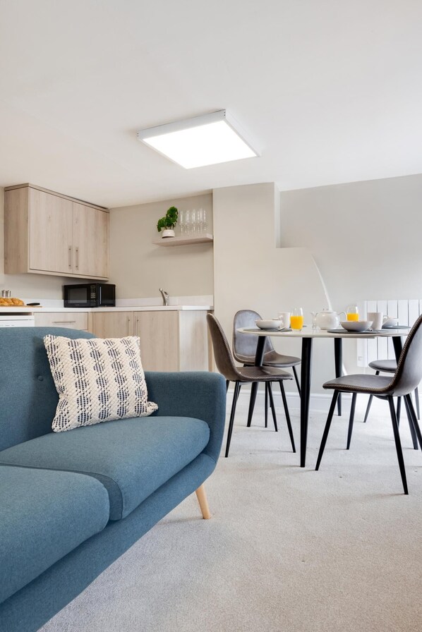 A modern living room with a blue sofa, a dining table with chairs, a kitchen area with cabinets and a microwave, and a ceiling light fixture. - 2 Bed in Holt (oc-2221) (Holt)