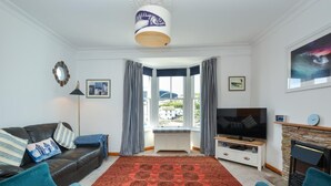 Een woonkamer met een bank, twee fauteuils, een televisie, een open haard en een groot raam met gordijnen.