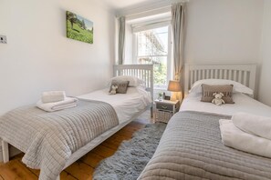 Ein Schlafzimmer mit zwei Betten, einem Fenster mit Vorhängen, einem Nachttisch mit Lampe und einem Teddybären auf einem der Betten.