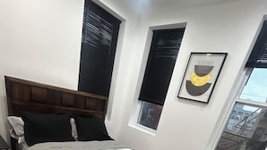 A bedroom with a large bed, wooden headboard, black and white bedding, a black throw pillow, a window with blinds, and a framed picture on the wall.