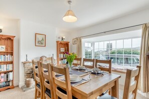 Un comedor con una mesa de madera preparada para cuatro personas, rodeada de sillas. Hay estanterías a ambos lados de la mesa, y una ventana con vistas a un coche y edificios.