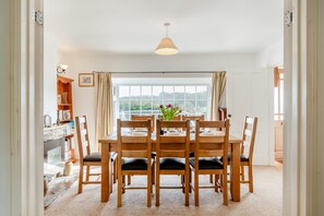 A dining area with a wooden table and chairs, a view through large windows, and a hanging light fixture. - 4 Bed in Stokenham (oc-i34021) (Stokenham)