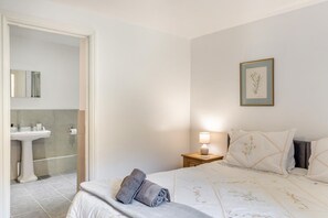 Um quarto com uma cama, uma mesa de cabeceira com abajur, uma ilustração botânica emoldurada na parede e um banheiro visível através de uma porta aberta.